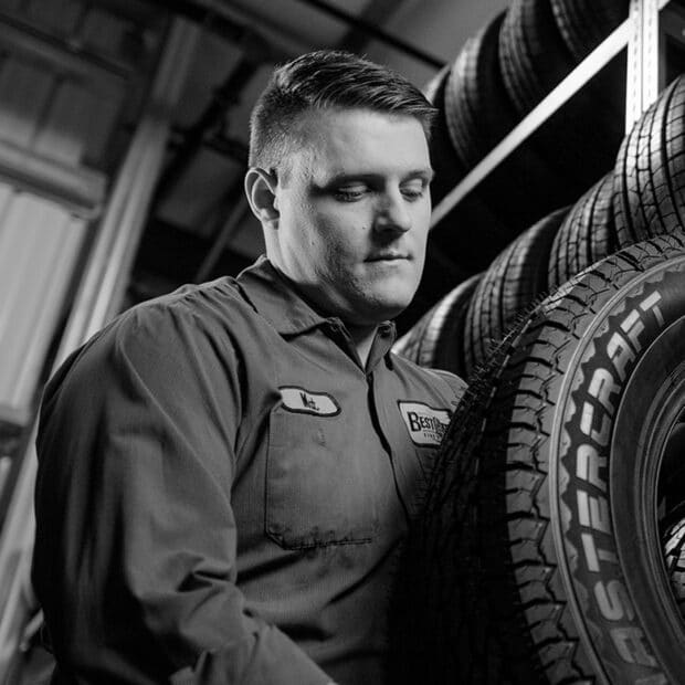 A mechanic in uniform inspects a tire in a garage with shelves of tires in the background. The scene has a focused and professional tone.