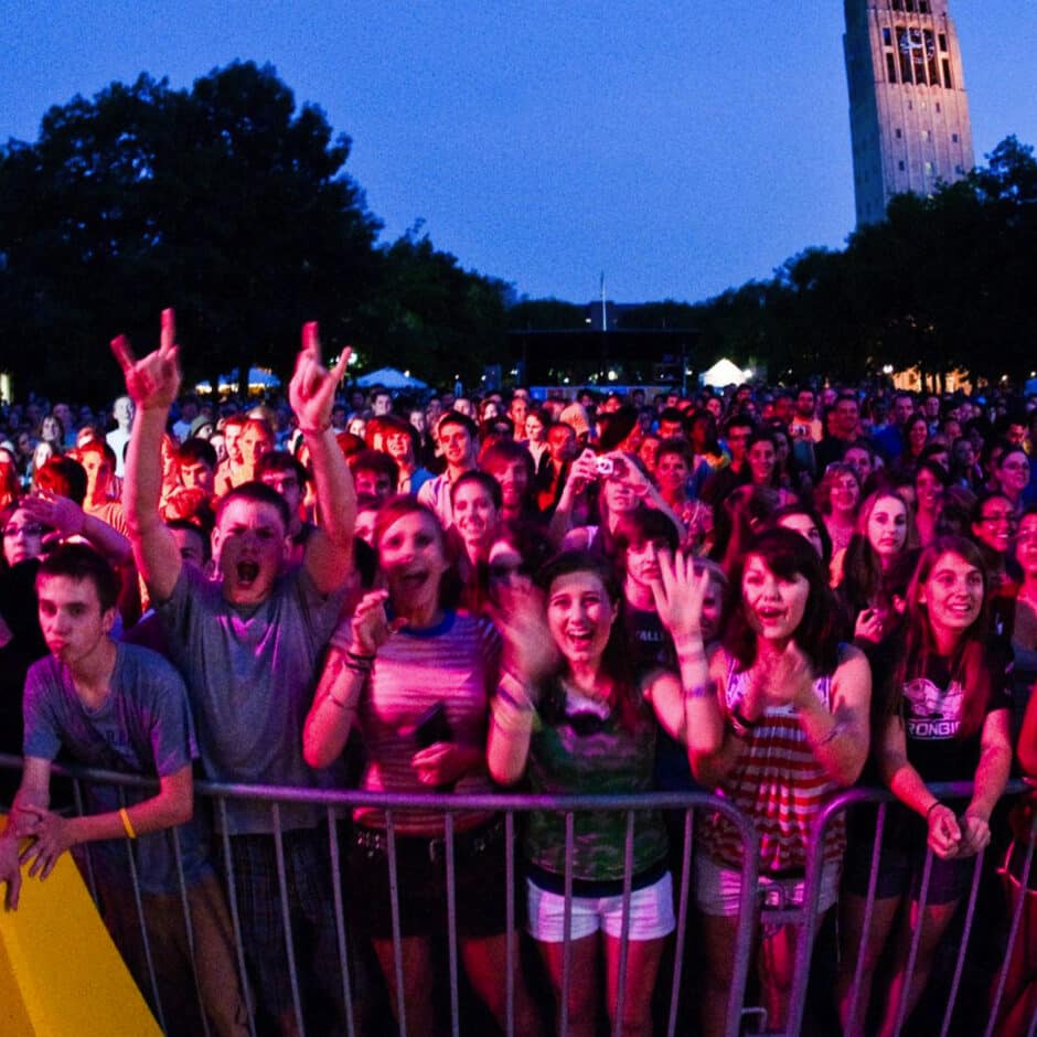A large, excited crowd of mostly young people stands behind a barrier at an outdoor concert during twilight. A tall clock tower is visible in the background.