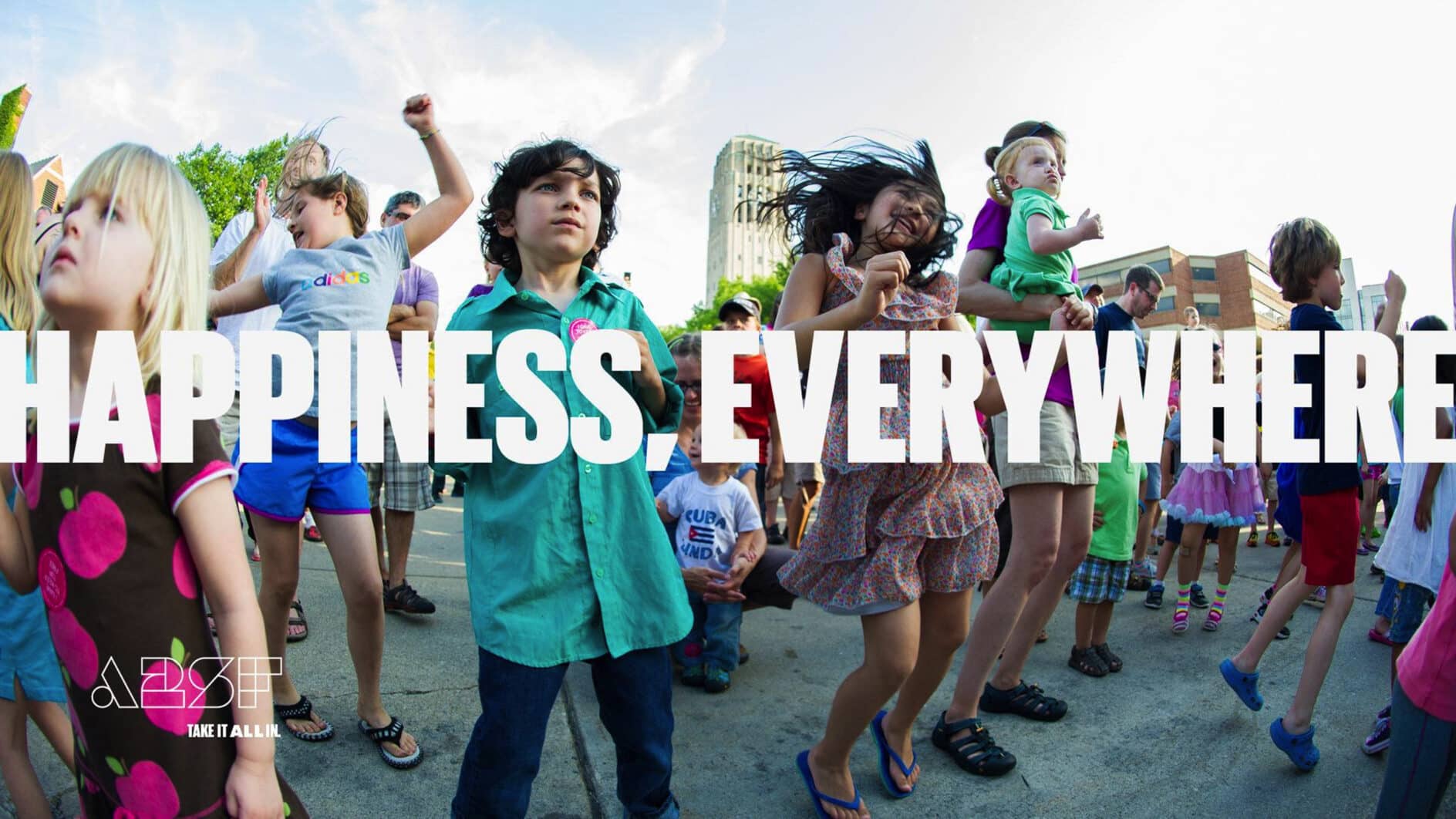 Children joyfully dance outdoors at a festival, exuding happiness. The text "Happiness Everywhere" overlays the scene, emphasizing the joyful mood.