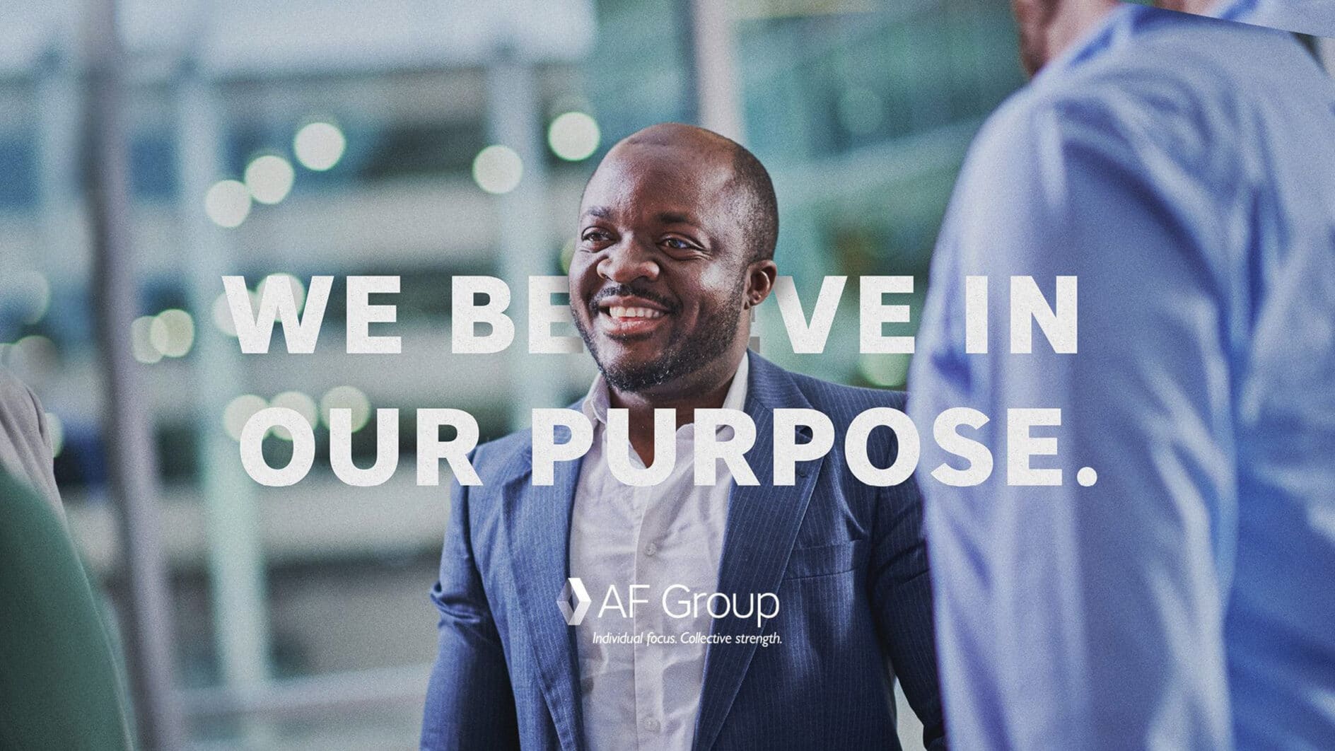 A man in a suit smiling and interacting with others, text overlay reads "We believe in our purpose." The background is a blurred office setting.