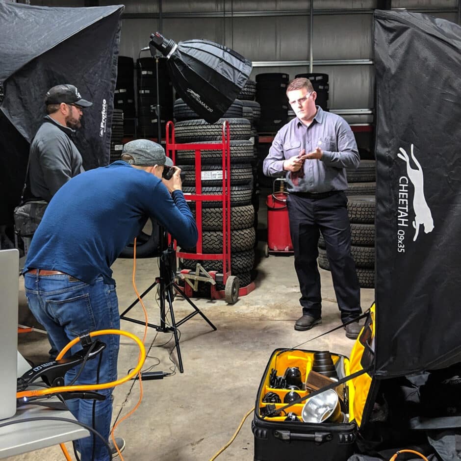 A photoshoot is underway in a garage with stacks of tires. A photographer in a blue shirt captures a man adjusting his shirt, surrounded by studio lighting and equipment. The scene conveys a focused, professional atmosphere.