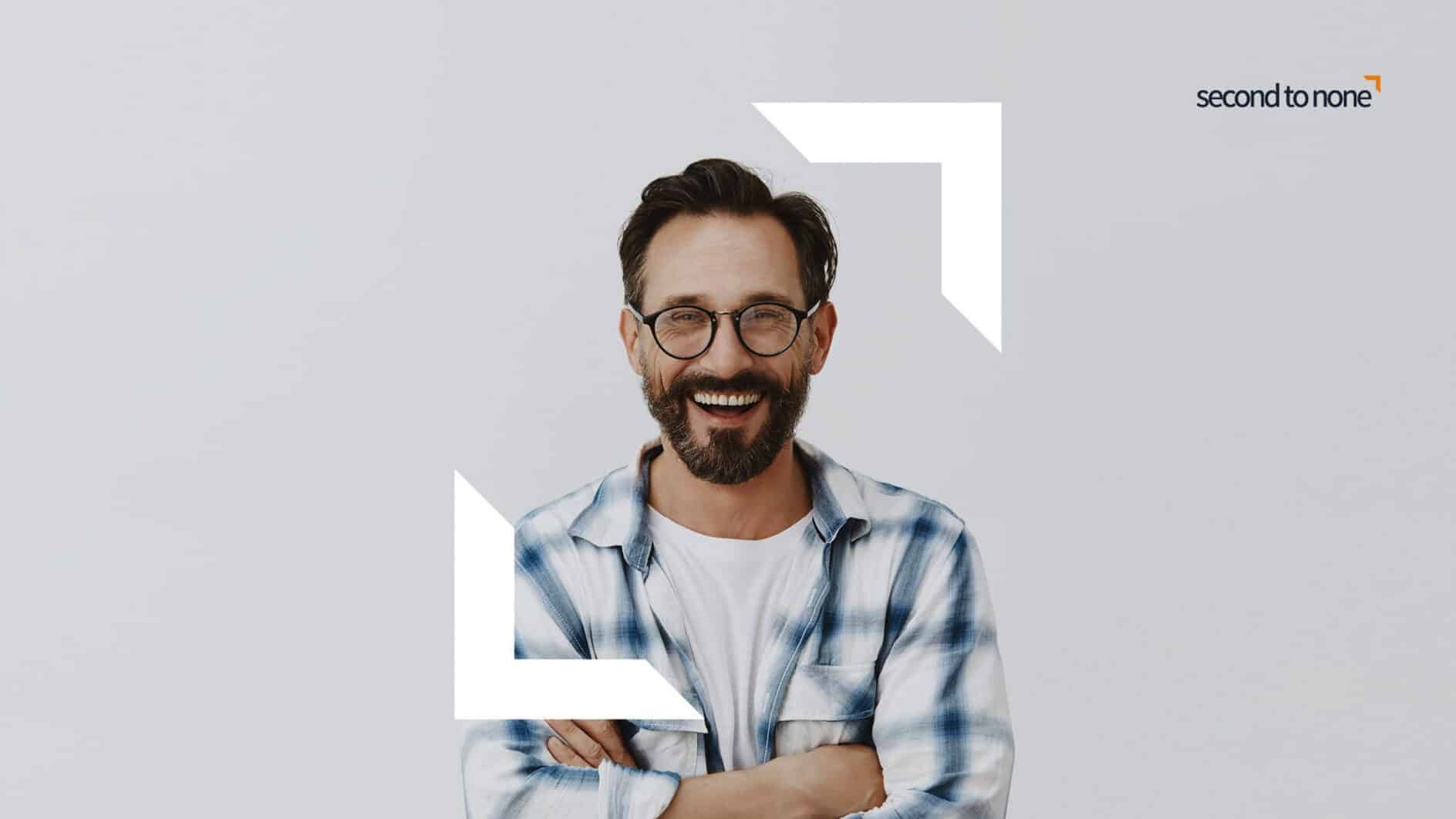 Smiling man with glasses and a beard, wearing a blue plaid shirt, stands with arms crossed. The background is light gray with the text "second to none."