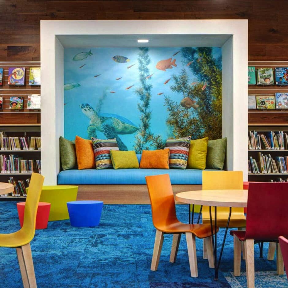 Colorful children's reading nook in a library, featuring a vivid ocean mural with a turtle, bright cushions, vibrant chairs, and surrounding bookshelves.