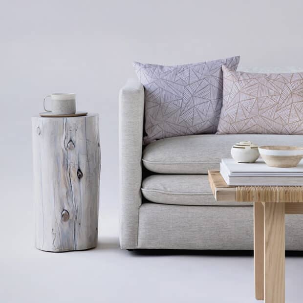A minimalist living space with a light gray sofa featuring geometric-patterned cushions. A rustic wooden side table holds a ceramic mug, adding a cozy touch.