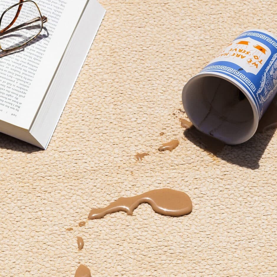 Spilled coffee from a tipped paper cup on a beige carpet, with a pair of glasses resting on an open book nearby. The scene conveys a mild sense of chaos.