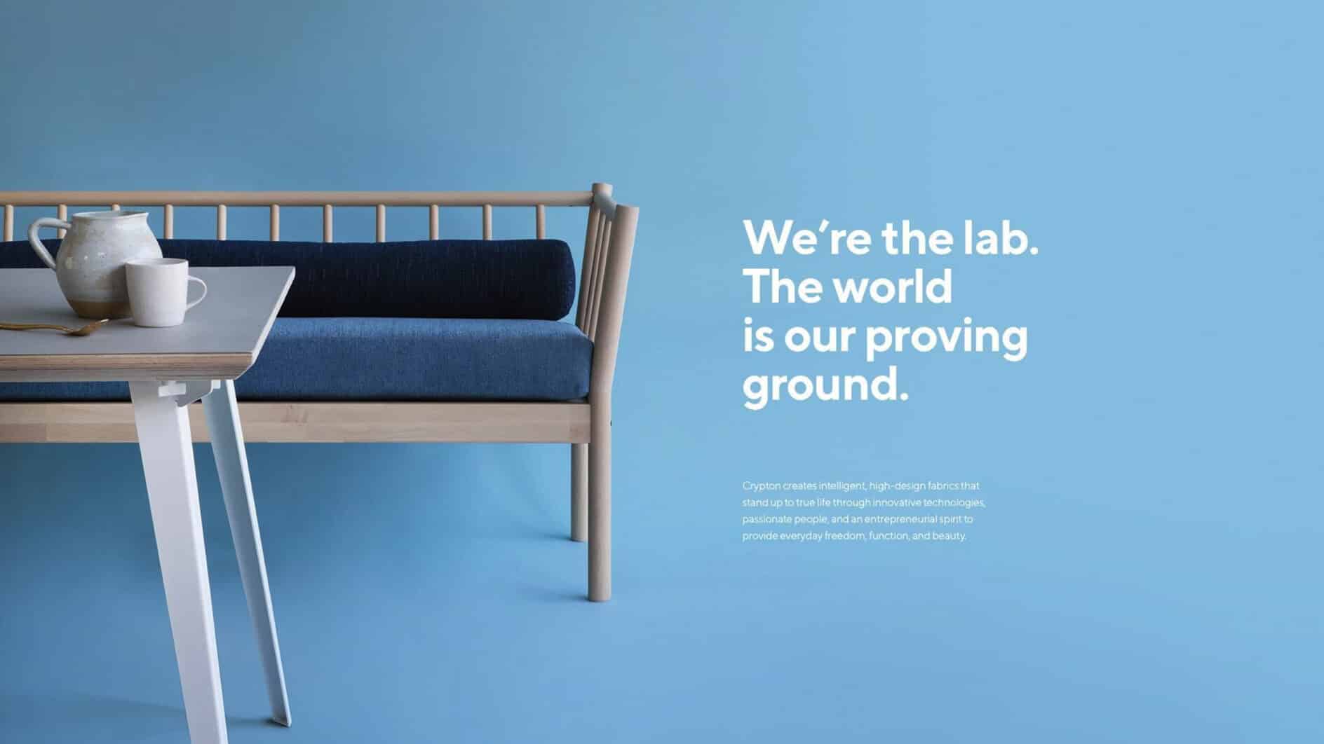 A modern wood-framed loveseat with blue cushions sits beside a table holding a teapot and cup. On a blue background, text reads, “We’re the lab. The world is our proving ground.”