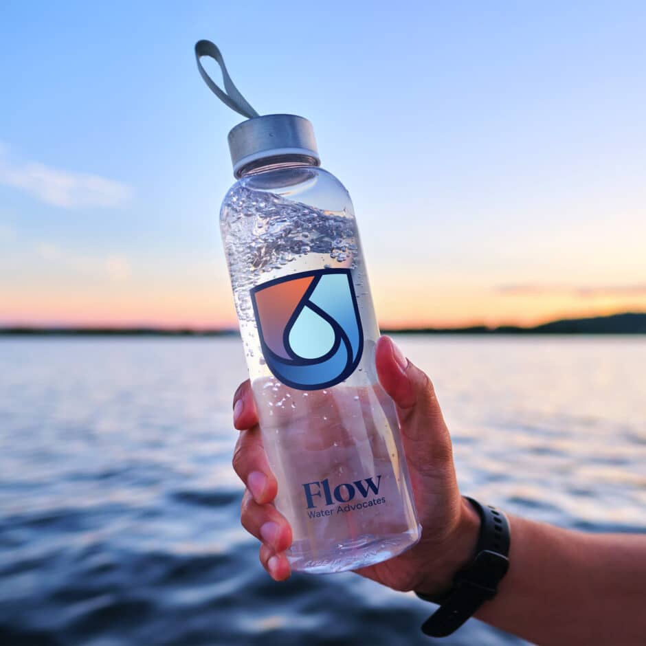 A hand holds a clear water bottle with a logo against a serene lake backdrop at sunset, with soft pink and orange hues in the sky.