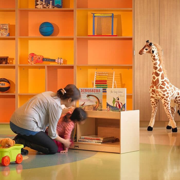 A woman helps a child in a colorful playroom with orange and yellow shelves holding toys and books. A toy giraffe stands nearby, creating a joyful atmosphere.