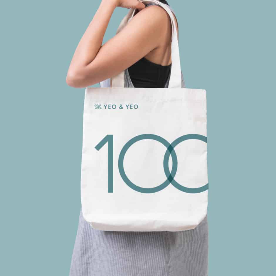 A person carries a white tote bag with the text "Yeo & Yeo" and "100" in teal. The background is a light blue-green, conveying a modern, minimalist tone.