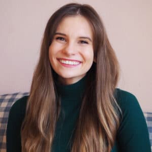 Smiling woman with long brown hair and a green turtleneck sits on a checkered couch. The background is a soft peach color, conveying warmth and friendliness.