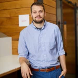 A man in a light blue shirt and blue pants leans against a white desk, smiling slightly. The background has warm wooden paneling, conveying a casual and professional tone.