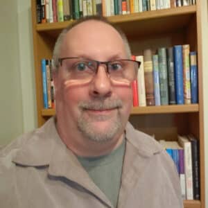 A person with glasses and a short beard smiles slightly in front of a bookshelf filled with colorful books, creating a warm and scholarly atmosphere.