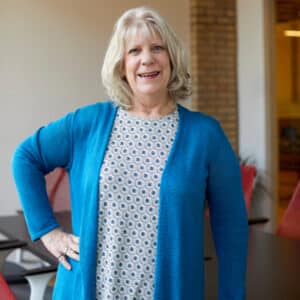 Woman with shoulder-length gray hair smiling warmly, wearing a blue cardigan and patterned blouse, standing in a conference room. Engaging and professional tone.