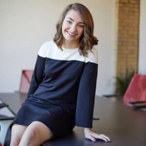 A smiling woman in a black-and-white dress sits casually on a table in a modern office. Red chairs and a brick wall suggest a professional setting.