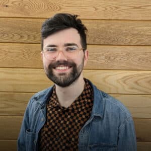 A man with glasses and a beard smiles gently, wearing a checkered shirt and denim jacket. He stands against a light wood-paneled wall.