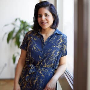 Smiling woman with dark hair in a floral blue dress leans against a windowsill with light streaming through. A leafy plant is in the blurred background.