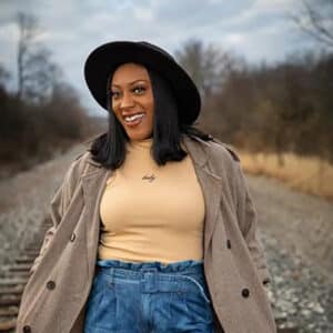 Smiling woman in a tan coat and black hat stands on a railroad track. Overcast sky and leafless trees create a serene, autumnal atmosphere.