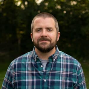 A man with short hair and a beard stands outdoors, wearing a blue plaid shirt. The background is blurred greenery, conveying a calm, relaxed mood.